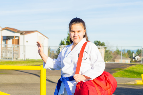 A young girl in a martial arts gi stands in front of a school fence.