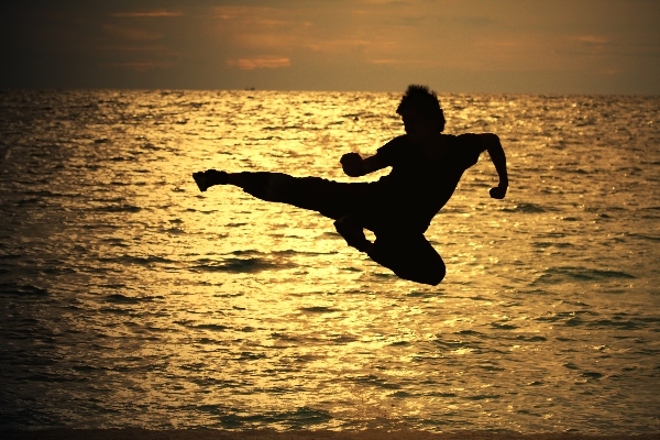 A Jeet Kune Do martial artist does a jumping side kick silhouetted by the sunset on a beach.