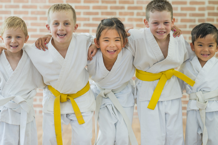 A group of kids in martial arts gis smile at the camera, holding each other as teammates.